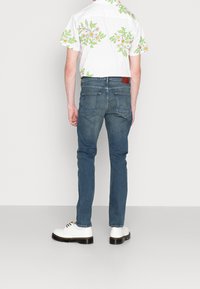 Bílá košile s krátkými rukávy a barevným květinovým potiskem, doplněná tmavě modrými džíny a bílými botami. Na džínách jsou viditelné zadní kapsy.