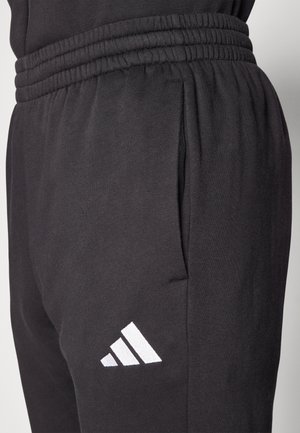 Svarta sportbyxor i mjukt tyg, med resårmidja, sidofickor och en vit Adidas-logga på vänster lår.