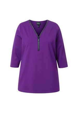 Tunic top viola con scollatura a V e chiusura con zip decorata con borchie. Presenta maniche tre quarti e una texture liscia.