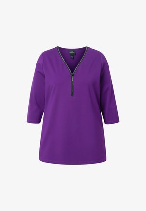 Tunic top viola con scollatura a V e chiusura con zip decorata con borchie. Presenta maniche tre quarti e una texture liscia.