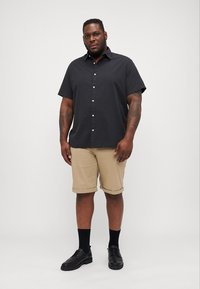 Chemise noire à manches courtes avec boutons, shorts beige, chaussures noires et chaussettes sombres. Design sobre, tissu léger et coupe moderne.