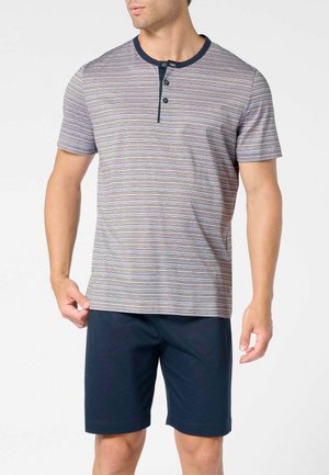 Uomo che indossa una camicia a maniche corte a righe con bottoni e pantaloncini scuri al ginocchio, su sfondo neutro.