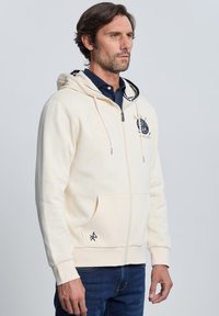 Sudadera con capucha color beige claro con cremallera, bolsillo tipo canguro, detalles bordados y capucha con cordón. Puños y dobladillo acanalados; textura suave.