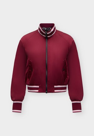 Kvinde med langt mørkt hår iført en rød bomberjakke, blå jeans, sorte sneakers, der holder en sort taske og står foran en ensfarvet hvid baggrund.