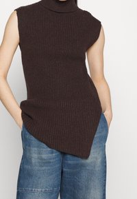 Turtleneck ärmelloses Top aus dunkelbraunem Rippstrick mit asymmetrischem Saum. Kombiniert mit hellblauen, weiten Jeansshorts.