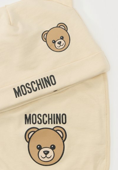 Μπεζ ύφασμα με δύο χαμογελαστά πρόσωπα αρκουδάκια και μαύρο κείμενο "MOSCHINO" τυπωμένο πάνω από κάθε αρκουδάκι.