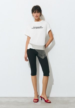 Ženska stoji ob beli steni, nosi belo majico s črnim napisom, črne kratke hlače, srebrn verižni pas in rdeče sandale z visokim petam.