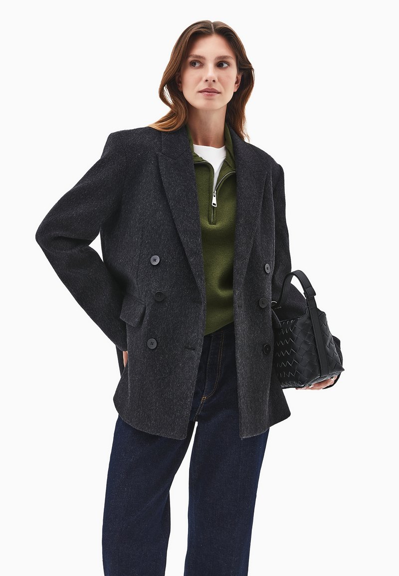 Tummanharmaa, kaksirivinen bleiseri, jossa on viistot läpät, yhdistetty vihreän vetoketjupullon kanssa ja pidetään teksturoitua mustaa laukkua.