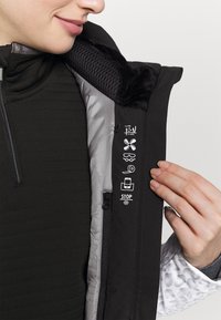 Personne tenant une veste noire ouverte montrant la doublure intérieure avec des icônes indiquant des caractéristiques étanches, respirantes, ventilées et isolantes.
