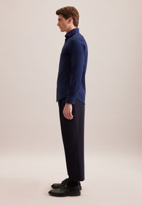 Navyblauwe overhemden met lange mouwen, slim fit en subtiele textuur, gecombineerd met donkere broeken en zwarte schoenen. Zijaanzicht.