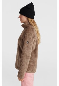 Bruine fleece pullover met hoge kraag, gestructureerd oppervlak, zijnaadzak en logo-opdruk op de linkermouw. Zwarte beanie op het hoofd.