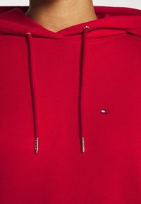 Rode hoodie met een trekkoord met metalen uiteinden en een klein logo label in de hoek. De stof lijkt glad en zacht te zijn.