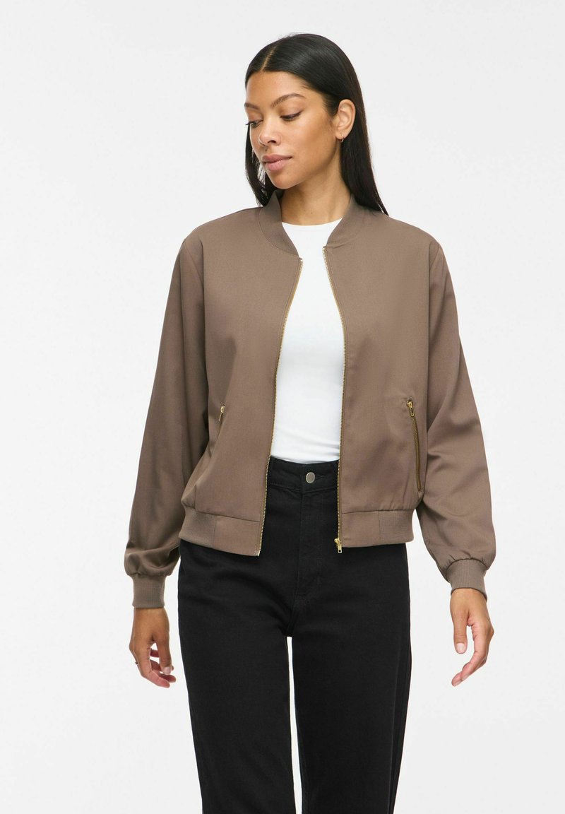 Femme portant une veste bomber marron zippée par-dessus un haut blanc et un pantalon noir, debout devant un fond blanc uni.