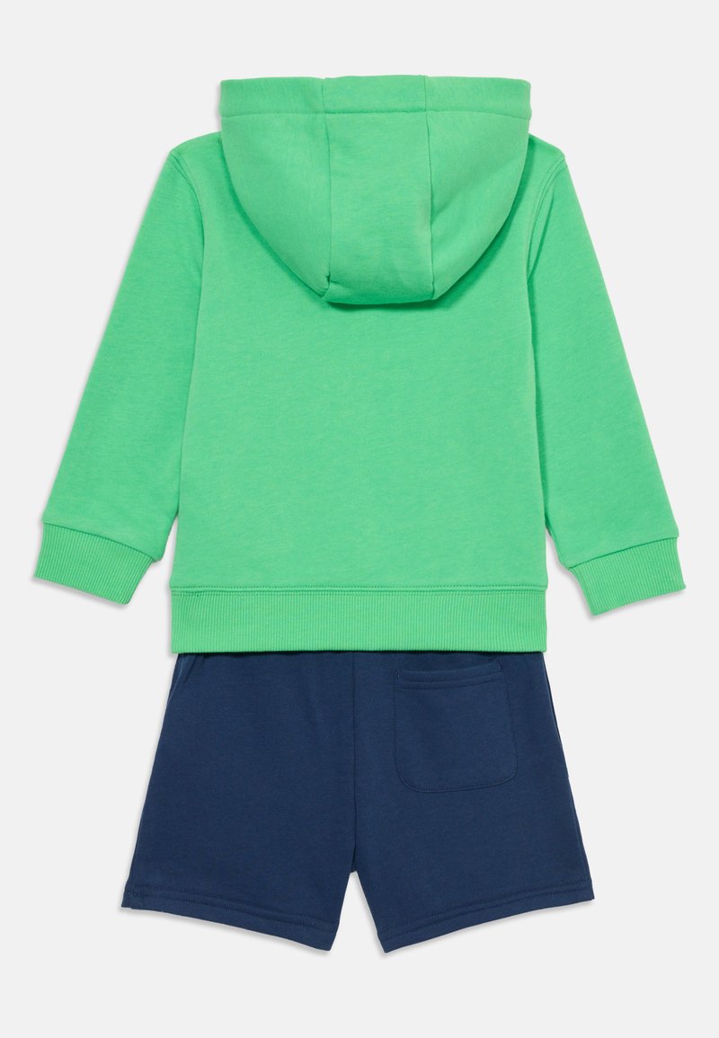 Felpa in cotone verde con cappuccio e polsini a costine abbinata a pantaloni shorts blu navy con tasca sul retro. Outfit casual con un design semplice.