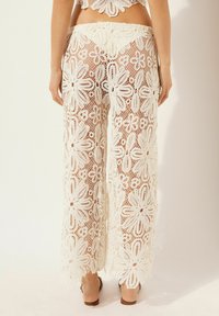 Pantalones de ganchillo blancos acampanados con patrones florales, diseño de tejido abierto, forma de pierna ancha y una cinturilla alta. Destacados por detalles delicados.
