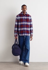 Plaid shirt in red, blue, and white with button closure, paired with blue denim pants and white shoes, carrying a round navy bag.