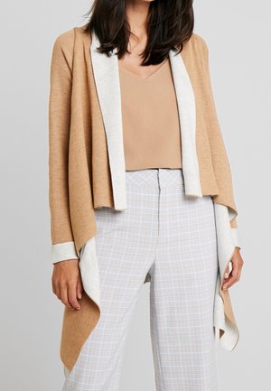 Cardigan leggero e aperto di colore tan con colletto e polsini color crema. Texture morbida, design a strati, indossato sopra una maglia beige e pantaloni a quadri.