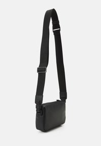 Bolso cruzado negro y rectangular hecho de piel suave con una correa texturizada y hebilla ajustable. Cuenta con una abertura superior con cremallera.