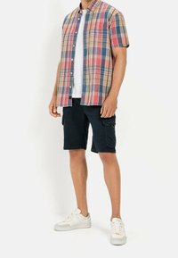 Kortærmet ternet skjorte i rød, blå og beige, parret med sorte cargo shorts. Hvid T-shirt under og hvide sneakers.