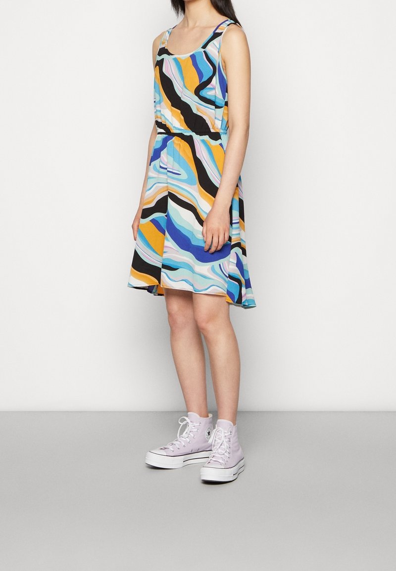 Barevné bezramínkové šaty mají vlnité vzory v modré, oranžové a černé barvě, s přiléhavým pasem a rozšířenou sukní. Na sobě má pastelově fialové tenisky.