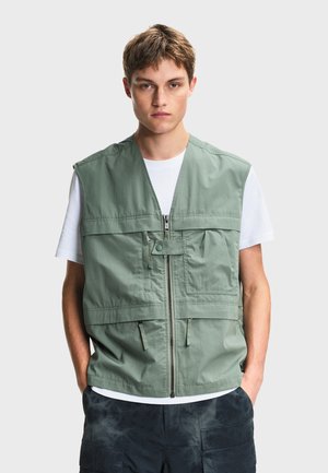 Jeune homme portant un gilet utilitaire vert clair avec plusieurs poches par-dessus un t-shirt blanc et un pantalon cargo foncé, debout les mains dans les poches.