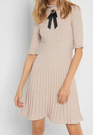 Femme portant une robe en maille côtelée rose clair avec des manches aux coudes et un nœud noir avec une boucle dorée au niveau de l'encolure.