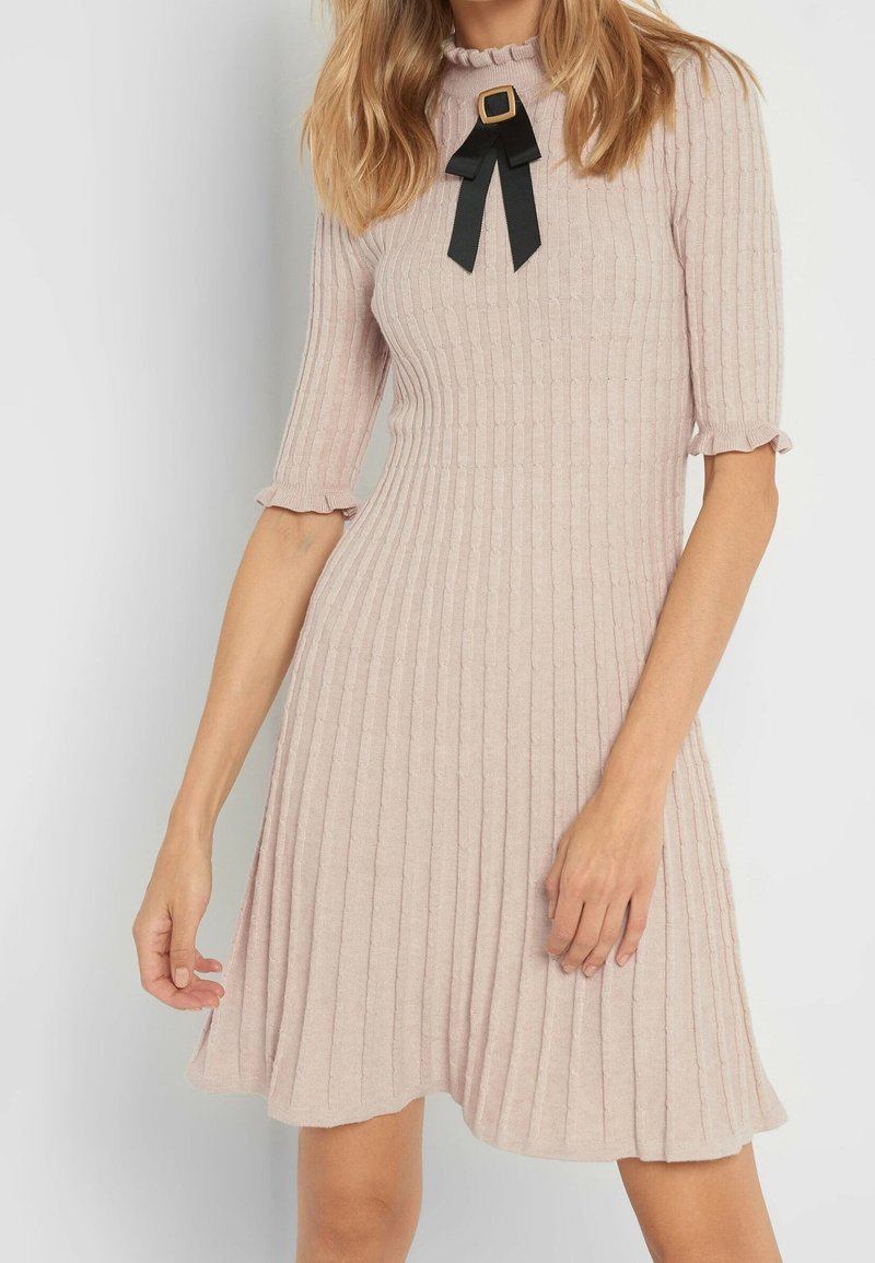 Femme portant une robe en maille côtelée rose clair avec des manches aux coudes et un nœud noir avec une boucle dorée au niveau de l'encolure.