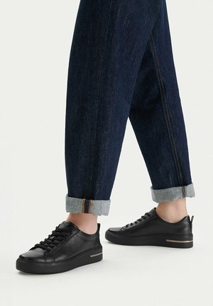 Persona que lleva vaqueros oscuros con puños y zapatillas negras de cuero bajas con cordones negros y una franja marrón en la suela.