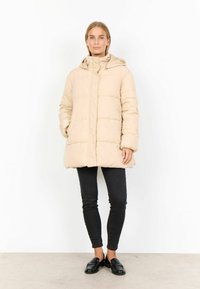 Abrigo de plumas beige con capucha, que presenta un diseño acolchado, cierre de cremallera frontal y dos bolsillos laterales, combinado con pantalones negros ajustados.