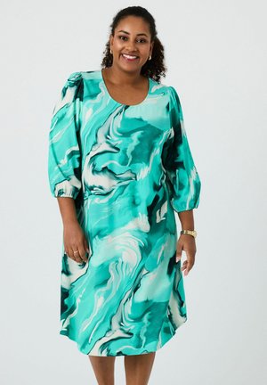 Woman smiling wearing a teal and white marbled patterned dress with puffed three-quarter sleeves and gold jewelry against a plain background.