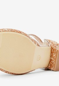 Glitzernder roségoldener Sandale mit einer strukturierten Gummisohle, einer breiten Riemen und einem glatten, breiten Blockabsatz. Die Unterseite zeigt Rillen.
