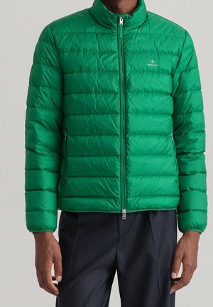 Man wearing a green quilted zip-up jacket with long sleeves and zippered side pockets, paired with dark pinstriped pants.