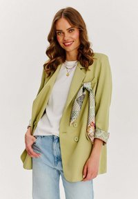 Jasnozielony blezer o luźnym kroju, z wzorzystym detalem na kieszeni i podwiniętymi rękawami. Noszony na białej koszuli i dżinsach.