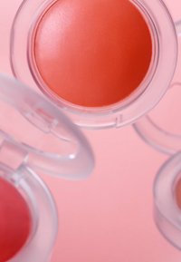 Blush créme rond dans une teinte corail clair, contenu dans un récipient transparent, entouré de deux autres blushs sur un fond rose doux.