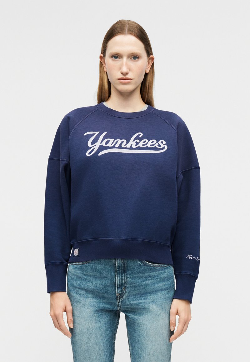 Γυναίκα που φοράει μπλε σκούρο φούτερ με την επιγραφή "Yamkees" σε λευκό γράμμα και ανοιχτό μπλε τζιν, στέκεται μπροστά σε ένα απλό ανοιχτό γκρι φόντο.