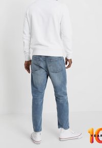 Witte trui, blauwe spijkerbroek en witte sneakers. Zichtbare achterzakken op de spijkerbroek, comfortabele pasvorm en enkellengte. Soepele textuur.