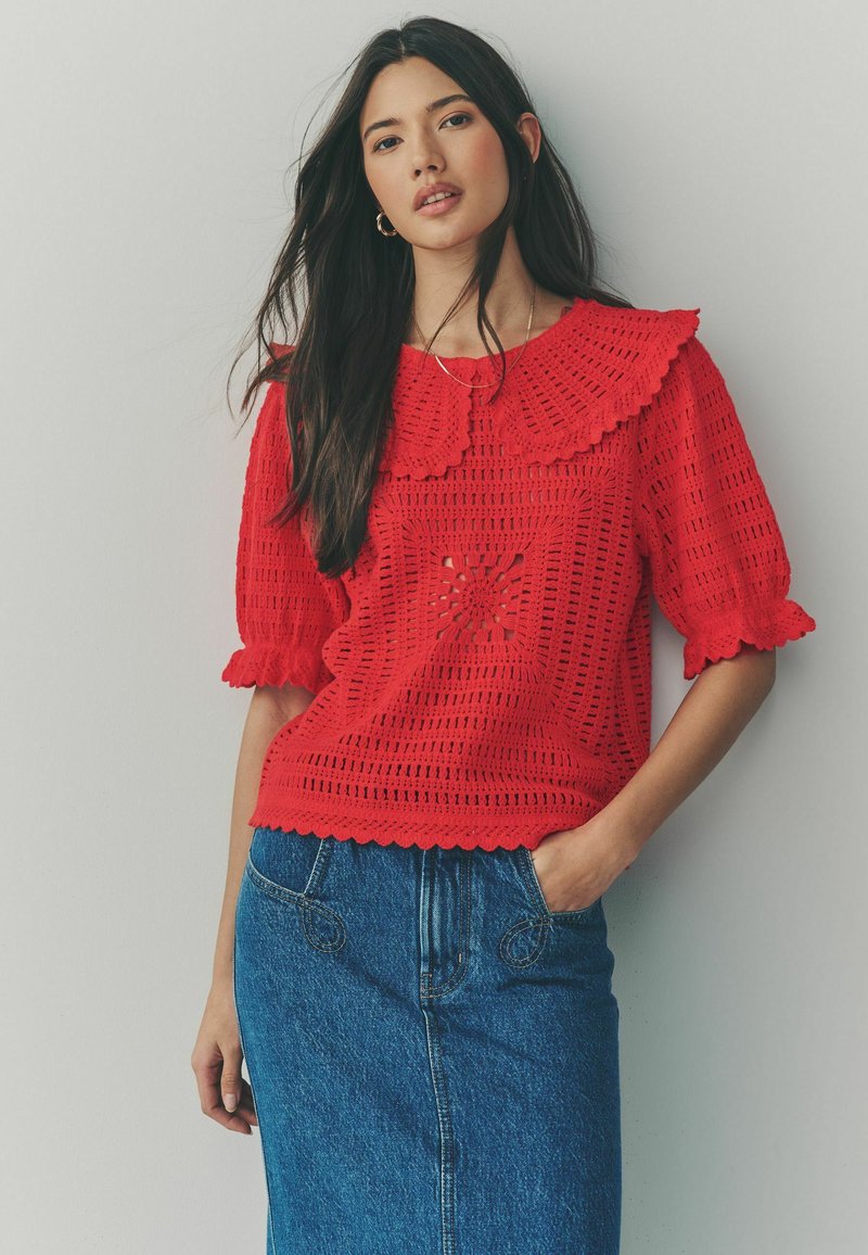 Haut en crochet rouge avec un col à volants, présentant un design en découpe florale. Assorti à une jupe en denim, mettant en valeur des motifs texturés.