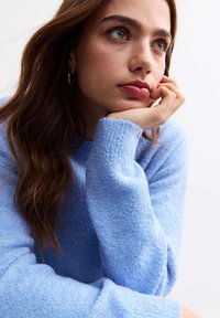 Maglione azzurro chiaro, con texture e maniche lunghe. La mano del modello poggia sul mento, parzialmente incorniciata da lunghi capelli castani. Aspetto morbido e accogliente.