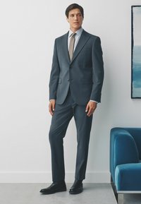 Dunkelblaues Anzug mit taillierter Passform, kombiniert mit einem hellblauen Hemd und einer gemusterten Krawatte. Schwarze Chelsea-Stiefel runden den Look ab.
