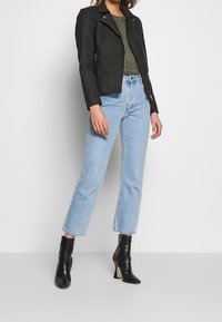 Musta tekonahkainen moottoripyöräjakku, jossa on uritettua yksityiskohtaa, yhdistettynä tiukkaan oliivinvihreään toppiin ja vaaleansiniisiin suoriin farkkuihin. Mustat korotettu nilkkabootsit.