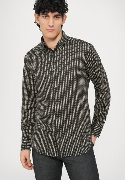 Juoda marškinėliai su baltu geometriniu raštu, ilgomis rankovėmis, užtrauktuku su mygtukais, smaigaliu apykakle ir kontrastingais mėlynais mygtukais. Dėvima su tamsiomis džinsais.