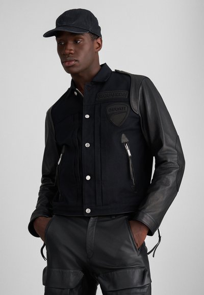 Chaqueta de mezclilla negra con mangas de cuero, que presenta un frente abotonado, dos bolsillos con cremallera y parches de marca en el pecho. Detalles de tela texturizada.