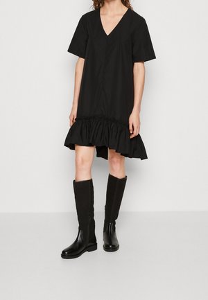 Persona con un vestido negro de manga corta con dobladillo con volantes y botas negras hasta la rodilla, de pie contra un fondo claro y liso.