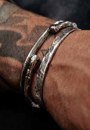 Zwei silberne Armbänder mit eingravierten Mustern an einem tätowierten Handgelenk mit dunkler Haut.