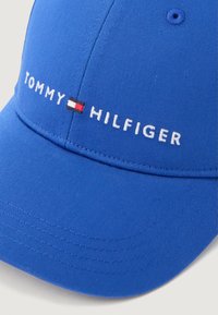Texturált anyagból készült kék sapka, fehér hímzett "TOMMY" és "HILFIGER" felirattal, valamint egy kis piros és fehér zászlóval díszítve.