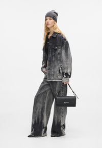 Μαύρο τζάκετ από τζιν σε oversized στυλ με κλίση χρώματος, συνδυασμένο με αντίστοιχες παντελόνες σε φαρδύ κόψιμο. Μια μαύρη τσάντα κρατιέται με καμπύλο λουράκι.