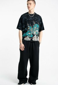 Młody mężczyzna w czarnej, oversize'owej koszulce z grafiką niebieskich smoków, luźnych czarnych spodniach, z srebrnym łańcuchem na szyi i bransoletkami, stojący na tle białym.