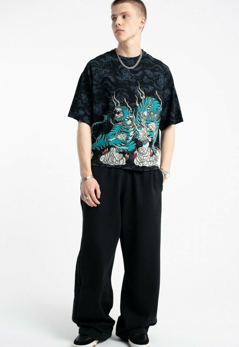 Młody mężczyzna w czarnej, oversize'owej koszulce z grafiką niebieskich smoków, luźnych czarnych spodniach, z srebrnym łańcuchem na szyi i bransoletkami, stojący na tle białym.