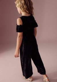 Zwarte fluwelen jumpsuit met off-shoulder ruffles, vloeiende wijde benen en textuurfinish. Zilveren sandalen maken de outfit compleet.