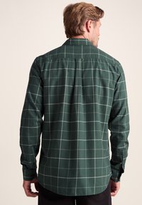 Green plaid button-up shirt with a relaxed fit, featuring a collar, long sleeves, and a curved hem. Light gray grid pattern throughout.